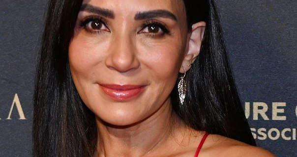 Marisol Nichols appears in front of a grey wall with pale gold writing on it. She wears her thick black hair straightened and loose. Marisol is seen in a pair of long gold leaf earrings, a gloss lipstick and black mascara.