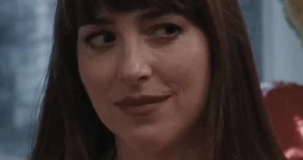 Dakota Johnson looks up and to the side. She wears a dark red matt lipstick with black mascara. Dakota stands in a large white wood window with red and yellow balloons to one side. Her light brown hair is long, thick and straightened with a fringe.