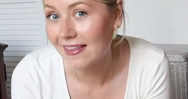 Meg Says sits on the floor of one of the bedrooms in her Surrey cottage. She has her long blonde hair tied back and up in a bun. Meg wears a white long sleeve top with gold earrings, a dark pink gloss lipstick and a thin gold necklace. She appears in front of a cream wicker storage basket and a white wooden wardrobe door.
