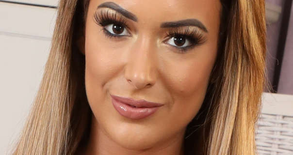 Lauren Louise stands in a purple and cream bedroom. She wears her light brown hair loose and down with blonde highlights. Laura appears in matt foundation, a glossy dark pink lipstick and lash curling foundation.