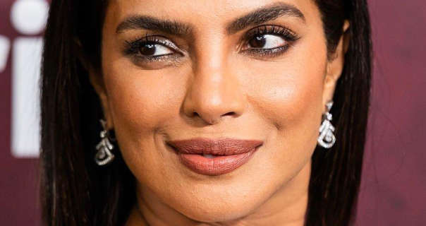 Priyanka Chopra looks to the side while smiling. She is seen in front of a maroon wall that has large white writing printed on it. Priyanka wears her long black hair straightened and down. She stars in a large of delicate silver earrings, black mascara, black eyeliner and a dark matt lipstick.