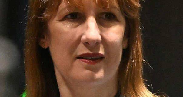 Rachel Reeves is seen with her hair loose and a little frizzy. She stands in a large dark studio room. Rachel wears a deep red lipstick with natural foundation and small earrings.
