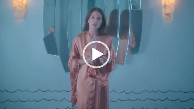 Julianne Moore stands in front of a cream wall and a large artisan mirror and two wall mounted orange lights. She wears a lace trim peach pink dressing gown. Julianne has her hair loose and pushed back.