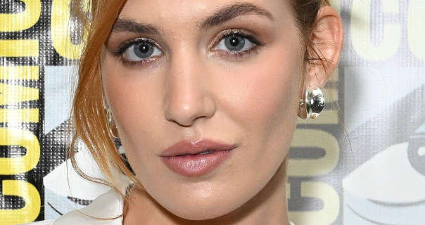 Sophie Nélisse is seen in front of a white, grey, black and yellow Comic-Con poster wall. She wears her hair tied back with strands pulled down to frame one side of her face. Sophie wears pink blush with a pair of large silver earrings, matt foundation, mascara and a dark pink lipstick.