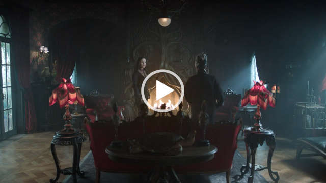 Catherine Zeta-Jones stands in front of a large lit open fireplace. She appears in an ornate gothic living room near a dark red sofa and two small display tables that have matching lamps on them. Catherine is seen in a black dress, heavy pale foundation and with her long black hair loose and pushed back.
