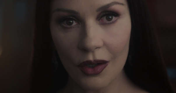 Catherine Zeta-Jones is seen in a very dark room. She has her black hair long and loose. Catherine wears a dark red lipstick with mascara and heavy pale foundation.