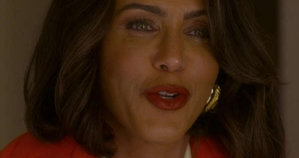 Nicole Ari Parker is seen with her light brown hair cut to shoulder length. She stands in a slightly dark apartment living room. Nicola is seen in a dark red lipstick, mascara, large gold earrings and a red jacket.