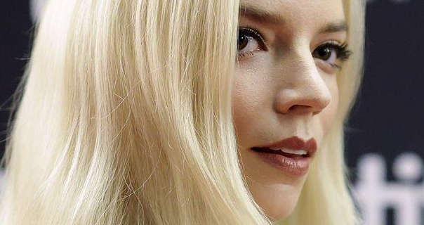 Anya Taylor-Joy is seen from the side with her long light blonde hair straightened and loose. She stands in front of a black wall with out of focus white text written on it. Anya wears mascara with a dark red lipstick and a red blush.