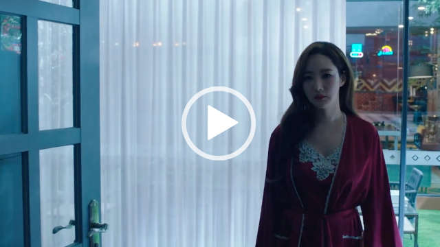 Park Min-young is seen in a dark pink silk and white lace dressing gown and nightdress. She walks through an open plan living room in the early evening. Park Min-young appears with her black hair loose and in gentle waves. She appears in front of a row of full height windows that have thin grey frames and sheer cream curtains.