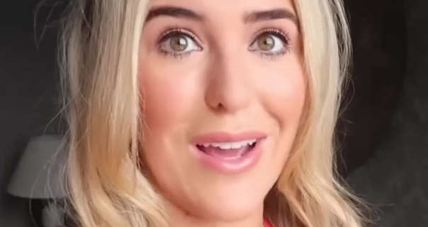Em Sheldon talks to her camera while vlogging. Her blonde hair is loose and straightened. She stands in a bedroom dressing area on her mum's wedding day near a small white lamp. She wears black mascara and black eyeliner with a light pink lipstick, foundation and soft pink blush.