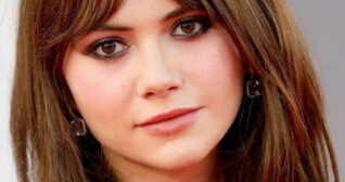 Emilia Jones appears in front of a light grey wall. She wears her thick brown hair straightened and loose. Emilia is seen in a pink lipstick, mascara, black eyeliner and a pair of square earrings.