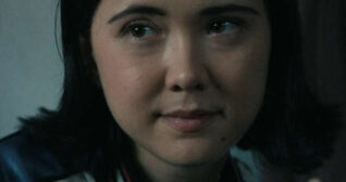 London Thor sits in a dark bedroom. She has her hair cut short to shoulder length. London wears a shiny white and blue bomber jacket with light blush, a pink lipstick and black mascara.