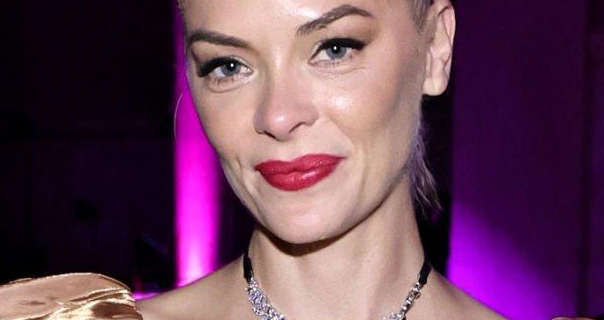Jaime King is seen in front of a black and vivid purple wall. She wears her blonde hair tied up and back. Jaime appears in a dark red lipstick, mascara and a silver gemstone necklace.