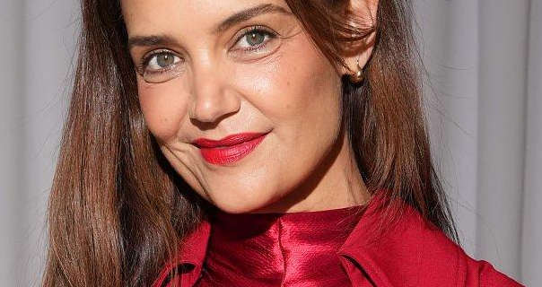 Katie Holmes stands in front of a drawn off white pleated curtain. She wears her thick light brown hair loose and with soft highlights. Katie wears a rich red lipstick with mascara, light blush, a red high neck dress and a red jacket.
