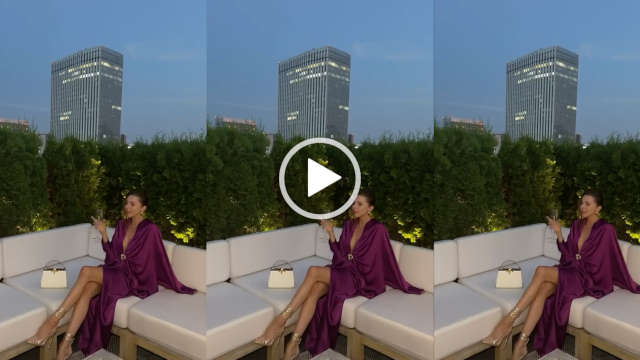 Laura Blair is seen sitting on a large cream and wood outdoor sofa. She wears a casual fit purple robe dress. Laura raises a wine glass and looks to one side. She appears in front of a green leafy hedge on a rooftop in Japan at dusk.