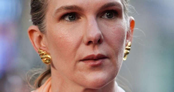 Lily Rabe stands outdoors with her blonde hair tied back in a ponytail. She is seen in a pair of large gold earrings and a peach colour dress. Lily wears a dark glossy lipstick with pink blush and black eyeliner.