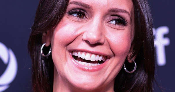 Nina Dobrev laughs and glances to one side. She stands in front of a navy wall that has white text and logos on it. Nina wears a glossy pink lipstick, black mascara and a pair of gold hoop earrings. She wears her long thick black hair in soft waves and loose.