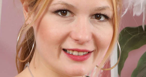 Freya H is seen with her hair tied back and in a blonde orange colour. She wears a pair of thin and large gold hoop earrings with black mascara and a dark pink lipstick. Freya sits in front of a dusty pink wall near white shelves a green leaf plant.