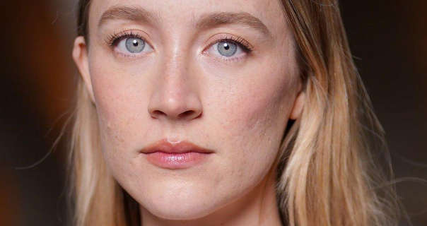 Saoirse Ronan wears her long blonde hair loose and in soft natural waves. She is seen in a light foundation, curled lashes and a glossy pale pink lipstick. Saoirse stands outdoors in front of a black and dark red out of focus backdrop.
