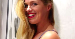 Lottie Lalay smiles and laughs while standing in front of a shower and a wall of cream bathroom tiles. She is seen with her long blonde hair loose and pull to one side. Lottie wears a bright red lipstick with a blue slip dress, mascara and no jewellery.