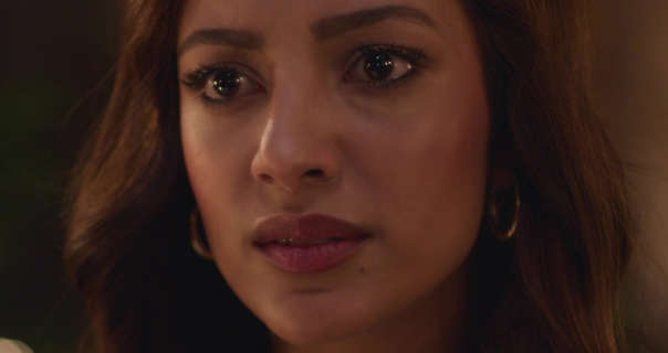 Sahher Bambba is looks forward with a serious expression and watery eyes. She stands in dark room with her light brown hair loose and in soft waves. Sahher wears a dark red matt lipstick with black mascara, black eyeliner and a pair of thin gold hoop earrings.