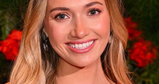 Tilly Keeper smiles with her long blonde hair loose and naturally styled. She wears a glossy pink lipstick with mascara and a pair of hanging dark gold circle design earrings. Tilly stands in front a green leaf and red rose wall.