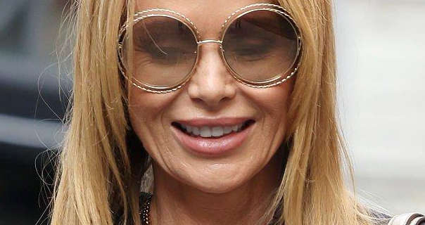 Amanda Holden walks outdoors through a London street on a slightly grey day. She has her long blonde hair straightened and loose. Amanda wears a pair of large circular rose gold wireframe sunglasses with a few large necklaces, foundation and a dark pink lipstick.