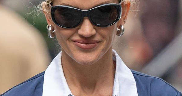 Ashley Roberts smiles and looks downwards. She is seen in a dark pink sheen lipstick, a pair of large gold earrings and black frame sunglasses. Ashley walks in London in a navy shirt that has a structured white collar. She has her long blonde hair tied back in a bun.
