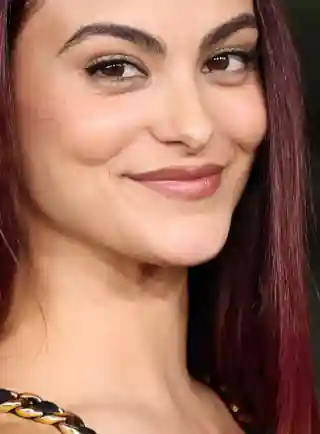 Camila Mendes smiles and looks into the camera. She is seen with red streaks through her straightened and loose black hair. Camila wears a black dress with flat foundation, mascara and a dark lipstick.