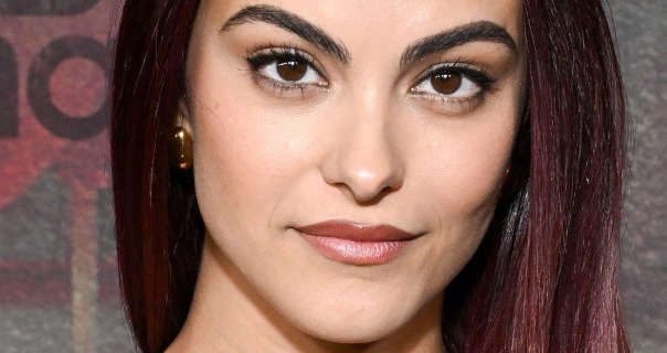 Camila Mendes is seen with red gradient highlights in her long black hair. She is seen in a matt foundation, mascara and a dark pink shiny lipstick. Camila stands in front of a grey and pastel red poster wall with her hair straightened, loose and pushed a little to one side.