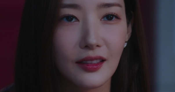 Park Min-young is seen in a dark room with her long black hair straightened and loose. She wears a light foundation with a little mascara and a dark red gloss lipstick. Park Min-young smirks as she looks directly ahead.