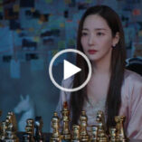Park Min-young looks forwards with a serious expression. She wears a light gold dressing gown with a gold and white lace nightdress. Park Min-young is seen in a dark room on a high back black chair. She sits in front of black and gold metal piece chess board with a large investigation board to one side.