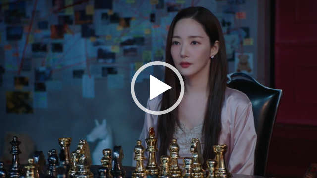 Park Min-young looks forwards with a serious expression. She wears a light gold dressing gown with a gold and white lace nightdress. Park Min-young is seen in a dark room on a high back black chair. She sits in front of black and gold metal piece chess board with a large investigation board to one side.