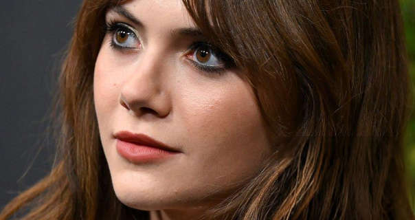 Emilia Jones appears in front of a dark grey wall. She has her thick light brown hair loose and naturally curled. Emila wears a light foundation with red blush, mascara, black eyeliner and a dark pink matt lipstick.