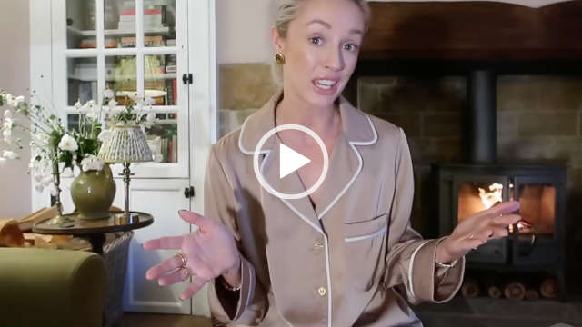 Josie Irons sits in front of a black metal log burner, a green sofa and a white full height cabinet at her home. She wears a pair of pale gold and white piping Holland Cooper pyjamas. Josie is seen with her blonde hair tied back. She appears in light makeup and a pair of large gold earrings.