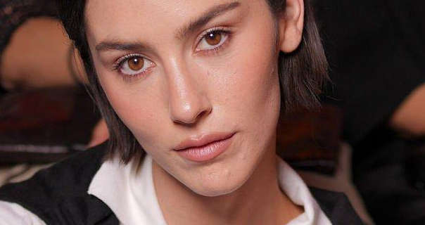 Gracie Abrams tilts her head a little to the side and looks straight into the camera. Her light brown hair is cut to shoulder length and is tucked back. Gracie wears a black sweater over her shoulders and with a white shirt. She appears in soft blush, black mascara and a dark pink sheen lipstick.