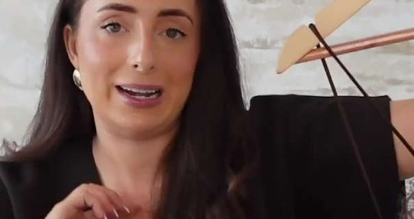 Hazel Maria Wood holds up a strappy top that is on a white and copper hanger. Hazel has her thick brown hair loose and straightened. She sits in front of a white brick effect wall. She wears foundation with a pair of gold earrings, black mascara and a dark pink lipstick.