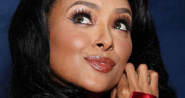 Kat Graham touches the side of her face with her hands. She glances upwards while standing in front of a dark blue poster wall. Kat wears a dark red gloss lipstick with foundation, a soft blush and heavy mascara. She wears her thick black hair down and slightly curled.