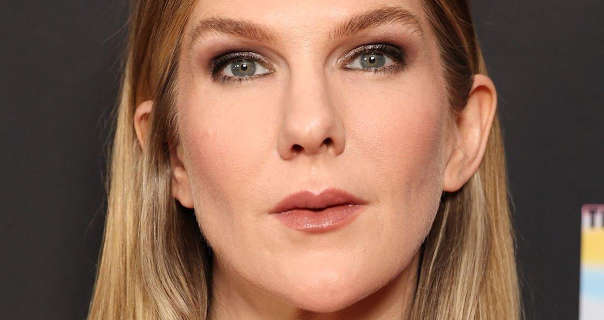 Lily Rabe is seen in front of a dark grey background with a colourful square logo just in frame. She has her long dark blonde hair loose and straightened. Lily is seen in a pink blush, black clumping mascara and a dark red gloss lipstick. She wears no jewellery.