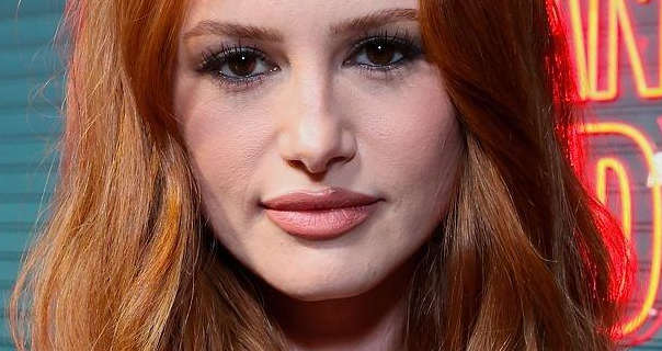 Madelaine Petsch stands in front of a blue metal sheet effect poster wall with neon wire frame writing behind her. She has her thick red hair loose and in soft waves. Madelaine wears a natural foundation with black mascara and a dark pink lipstick.