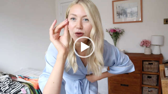 Meg Says holds up a hand and pinches her fingers together. She stands in front of a small wooden check of draws and a single bed in a cream and white bedroom at her home. Meg is seen with her long blonde hair in natural kinks, pulled forward and loose. She wears her light blue short dressing gown as she films a try on fashion haul.