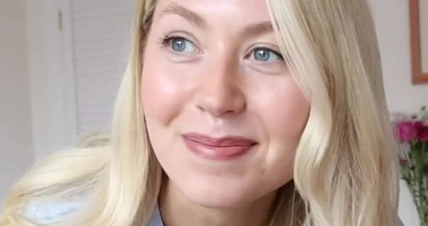 Meg Says leans forwards and looks to one side. She appears in her bedroom filming area in her cottage home. Meg has her long blonde hair loose and styled. She wears foundation, black mascara, soft blue and a dark red lipstick.