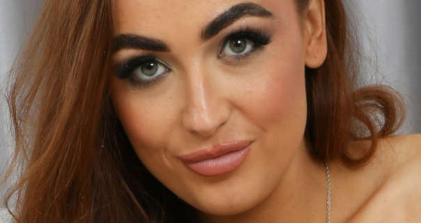 Blossom is seen with her thick light brown hair down and pushed a little to one side. She appears in front of a pair of drawn light grey living room curtains. Blossom wears heavy mascara with a dewy foundation, blush, light pink lipstick and a thin silver chain necklace.