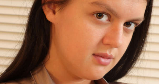 Kelsey is seen in front of a large closed shutter style blind in a yellow light. She wears her long black hair pushed back and loose. Kelsey is seen in a matt foundation, light pink lipstick and a gold short sleeve top.