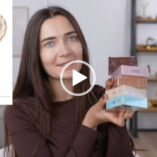 Roxxsaurus holds up five different blush palettes from Flower Knows. She sits in her tiny home filming studio set in front of a grey couch and wood ladder display shelves. Roxi wears a dark brown thin long sleeve top. She is seen with her long hair loose and straightened and with no makeup on.