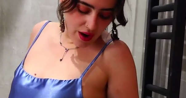 Valentina Boux stands in front of a cream and grey shower area. She wears her black hair tied up and with red streaks through it. Valentina looks downwards. She is seen in a pair of dangling earrings, a thin gold gemstone necklace, black mascara, blush and a matt red lipstick.