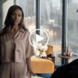 Nicole Beharie stand with her arms by her sides in front of a large living room window. She wears a pair of short sleeve gold pyjamas with her black hair loose and around her shoulders. Nicole appears in front of a light orange wood frame chair and small wooden table with a cityscape at daytime seen through the window.
