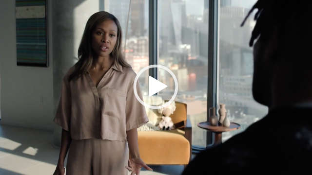 Nicole Beharie stand with her arms by her sides in front of a large living room window. She wears a pair of short sleeve gold pyjamas with her black hair loose and around her shoulders. Nicole appears in front of a light orange wood frame chair and small wooden table with a cityscape at daytime seen through the window.