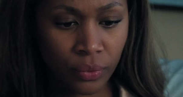 Nicole Beharie is seen on a dark cream and light blue bed. Her shoulder length black hair is a little curled and loose. She appears in a dark red lipstick and smoothing foundation.