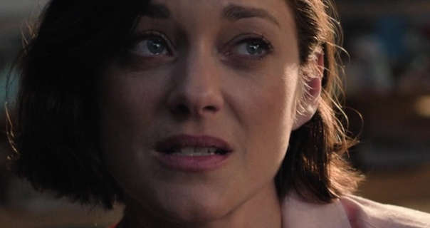 Marion Cotillard leans to one side. She is seen with her brown hair cut short and straightened. Marion appears in a pale pink shirt, blush, mascara and a dark pink lipstick. She sits in an open plan apartment kitchen area with sunlight hitting one side of her face.